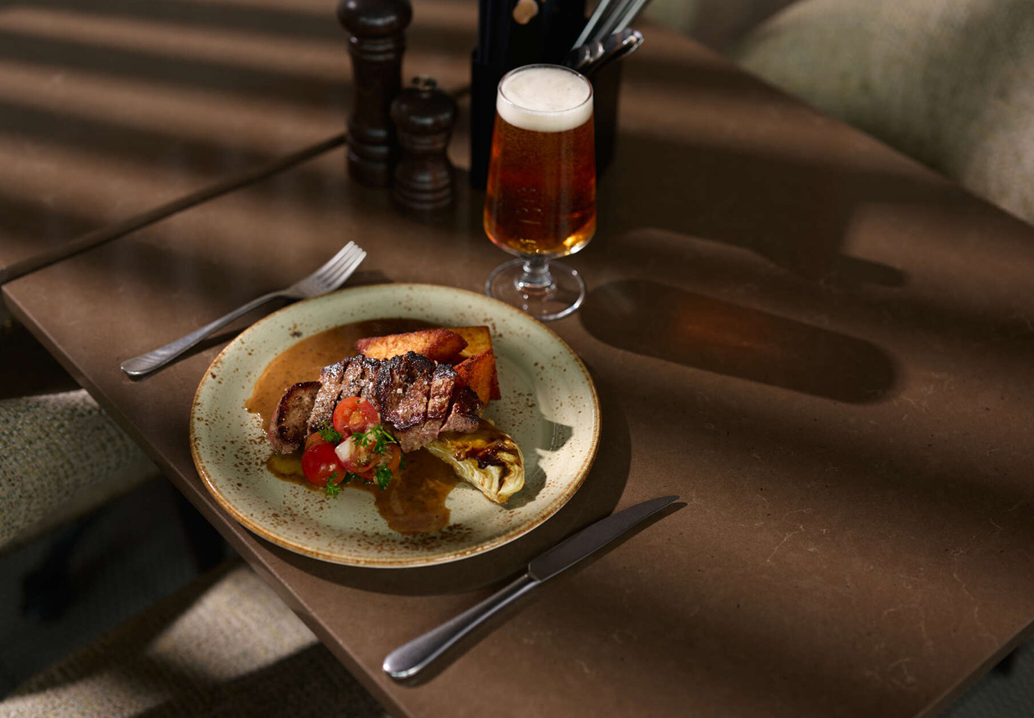 Tallrik med skivad biff, sås och tillbehör samt ett glas öl på bordet.